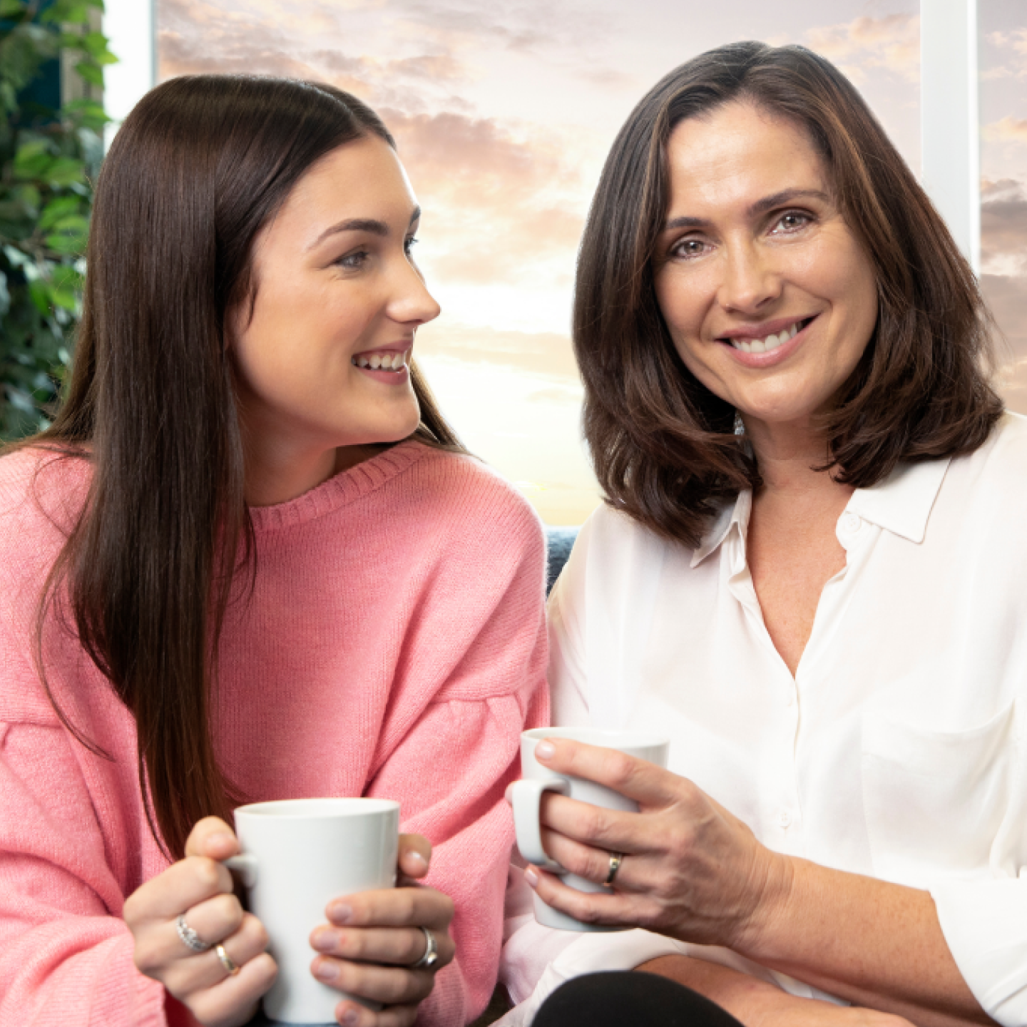 woman in a pink sweater holding a mug smiling at another smiling woman in a white shirt holding a mug