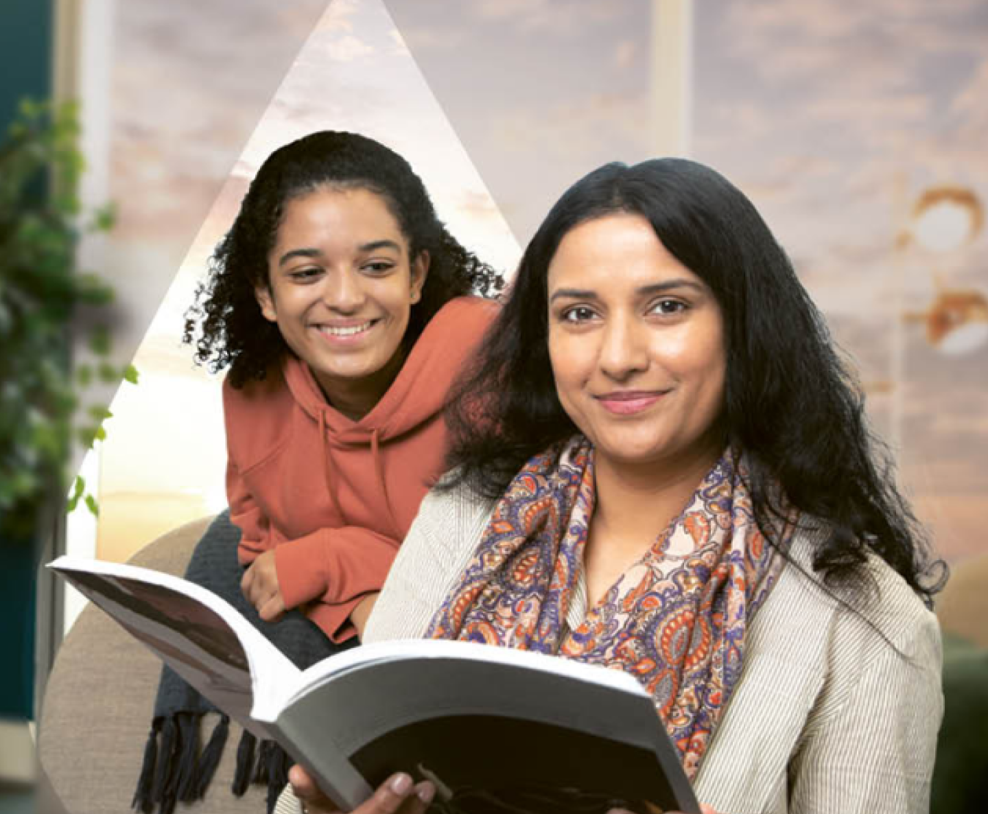 woman in a striped shirt and pattern scarf holding open a book and smiling at you with a curly haired woman in an orange sweater looking over her shoulder