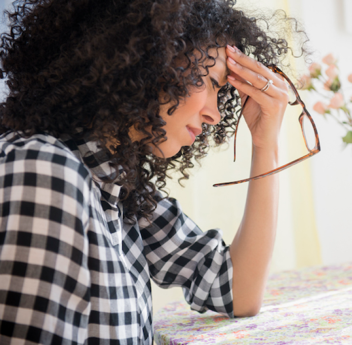 Woman with curly hair, holding glasses, touching her forehead in pain