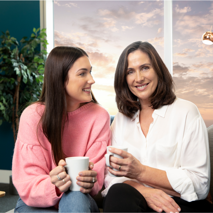 to kvinder sidder ved siden af hinanden og holder kaffekrus, mens de smiler
