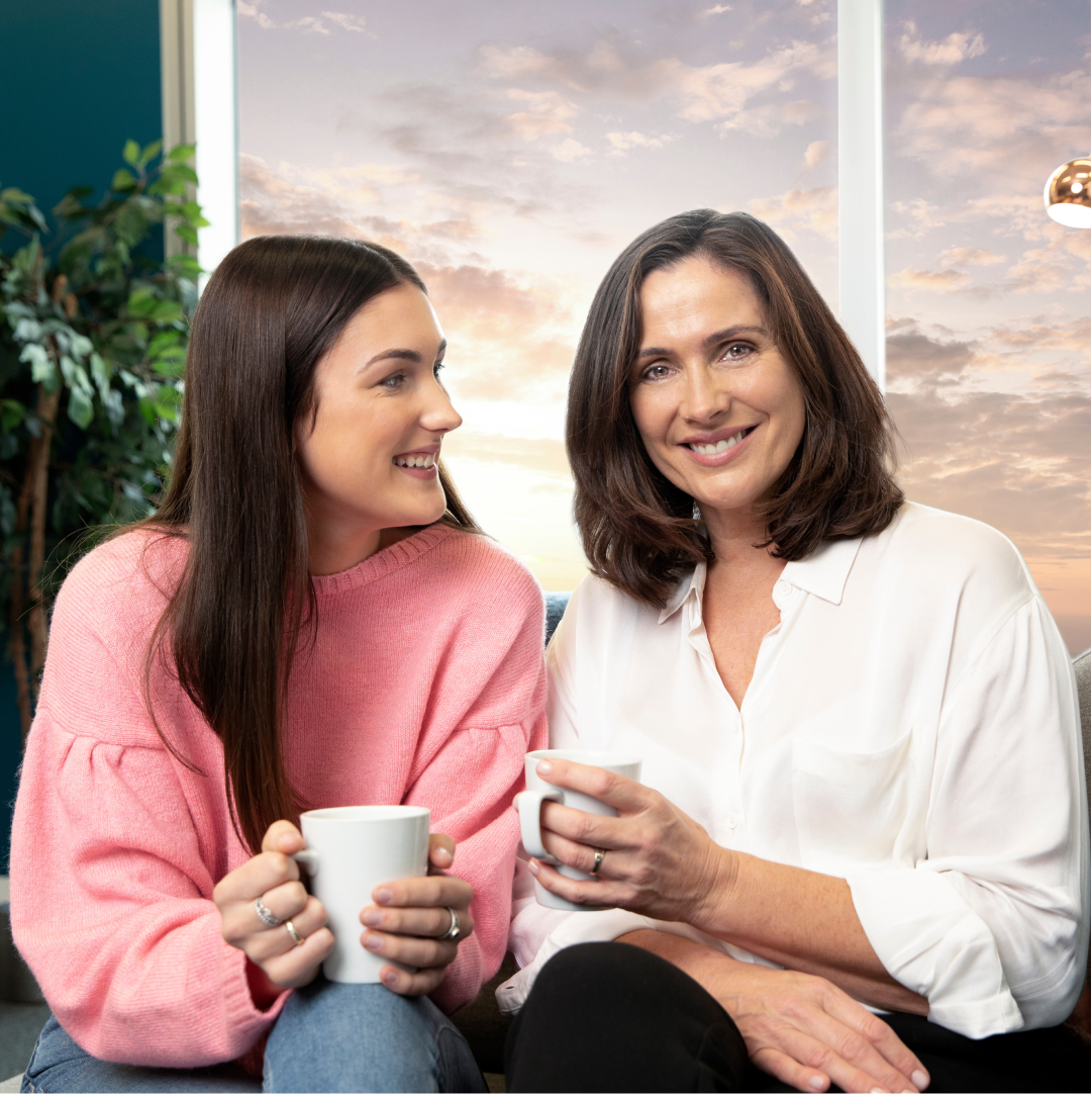 Dvije žene sjede jedna do druge držeći šalice kave dok se smiju.