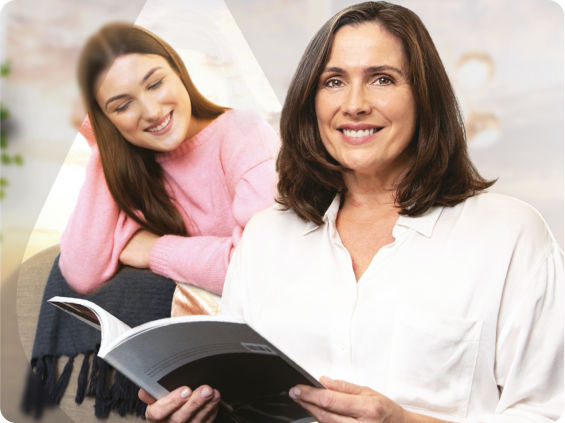 Žena u bijeloj košulji s gumbima drži knjigu dok se smije, a djevojka joj gleda preko ramena.