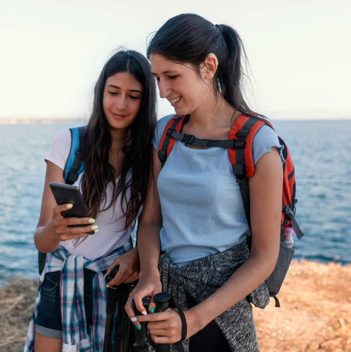 két fiatal nő néz egy telefont, hátizsákot viselnek, a háttérben természetes víz van