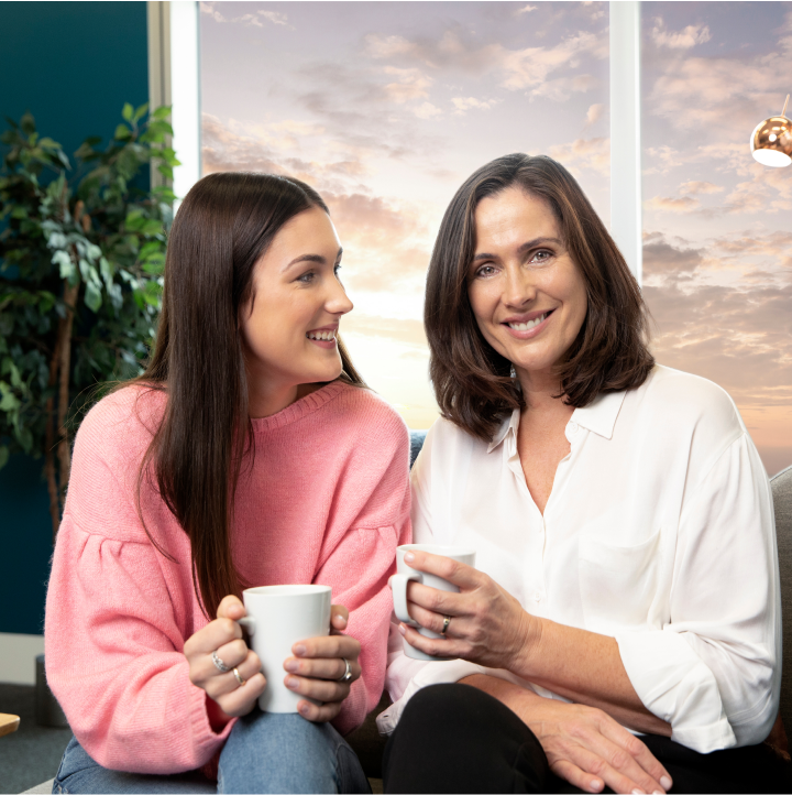 две жени седат една до друга држејќи шолји за кафе додека се смешкаат