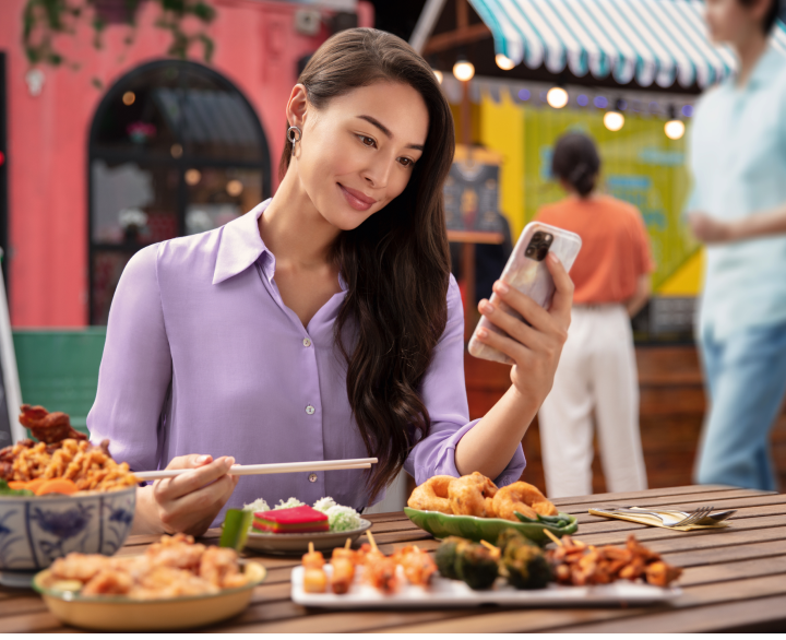 Woman sitting outside eating meal at restaurant holding chopsticks while looking at phone to take the Systane Stop&Stare Challenge