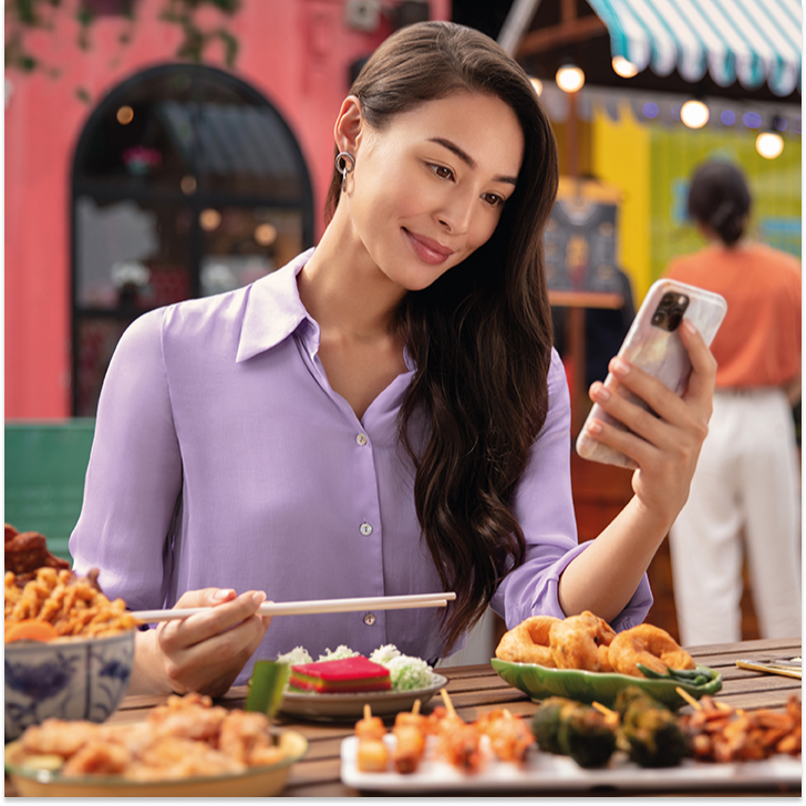 Woman sitting outside eating meal at restaurant holding chopsticks while looking at phone