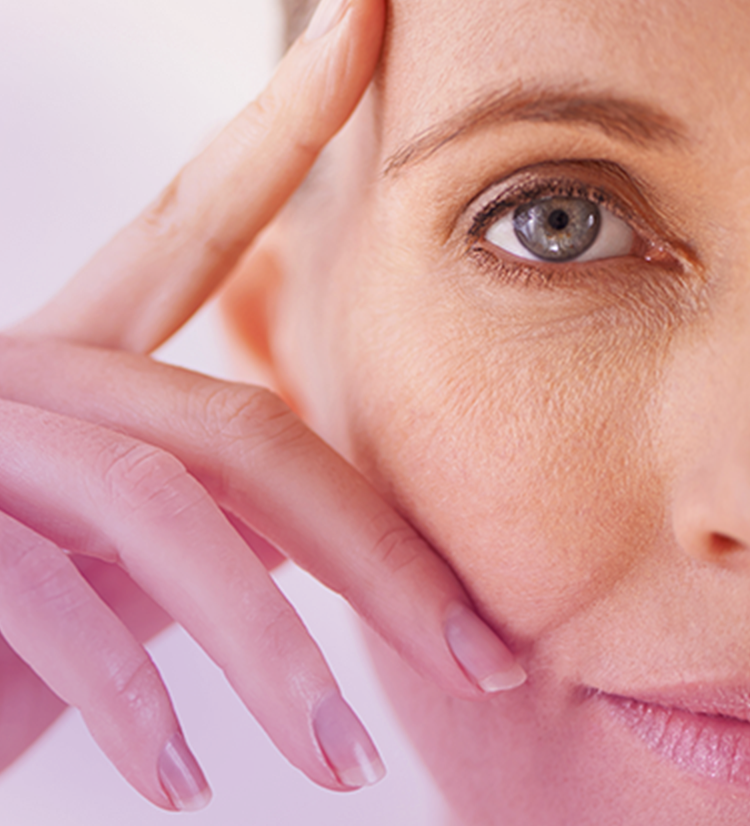 A close-up of a mature woman's face focusing on her right eye and the surrounding area. Her fingers are gently touching her temple, highlighting the fine lines and texture of her skin. The image emphasizes the natural aging process and the delicate area around the eyes.