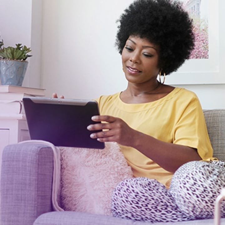 A woman with natural curly hair sits comfortably on a couch, holding a tablet researching dry eye. She is dressed casually in a yellow top and patterned pants, looking at the screen with a calm and focused expression.