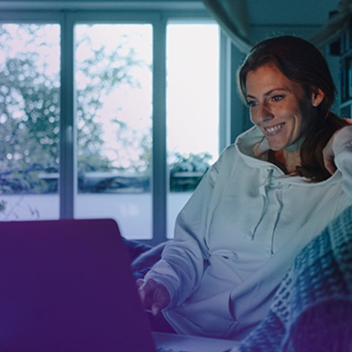 A woman wearing a white hoodie is sitting comfortably on a couch, smiling as she looks at her laptop screen
