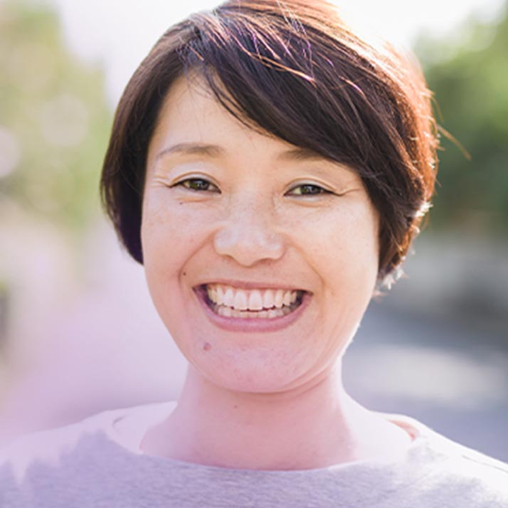 A close-up of a smiling woman with short, dark hair standing outside on a sunny day. The woman is dressed casually and her broad smile conveys a sense of happiness and positivity.