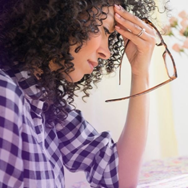 Woman with curly hair, holding glasses, touching her forehead in discomfort