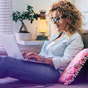 A woman with curly blonde hair and glasses sits comfortably on a couch, typing on her laptop. She is dressed casually in a light denim shirt and appears relaxed and focused.