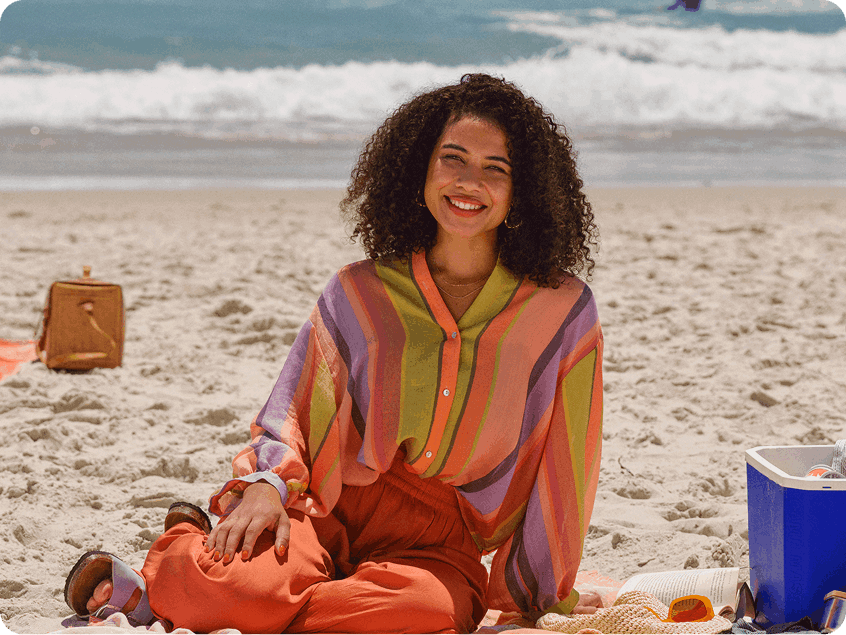 Pessoa sentada na areia da praia usando roupas coloridas, com o mar ao fundo e itens de praia ao redor.