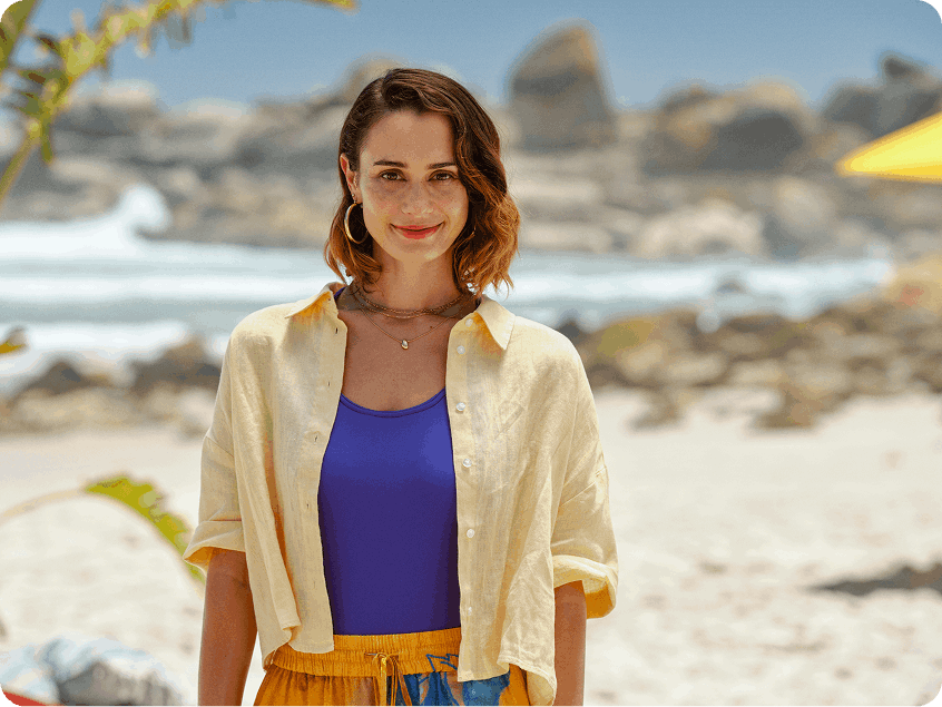 Pessoa em pé na praia usando camisa clara aberta sobre uma blusa azul, com o mar, rochas e vegetação ao fundo.