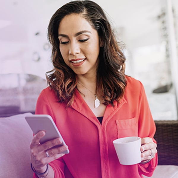 A woman with dark, wavy hair sits on a couch, looking at her smartphone while holding a white coffee mug in her other hand. She is dressed in a bright coral blouse and wears a delicate necklace.